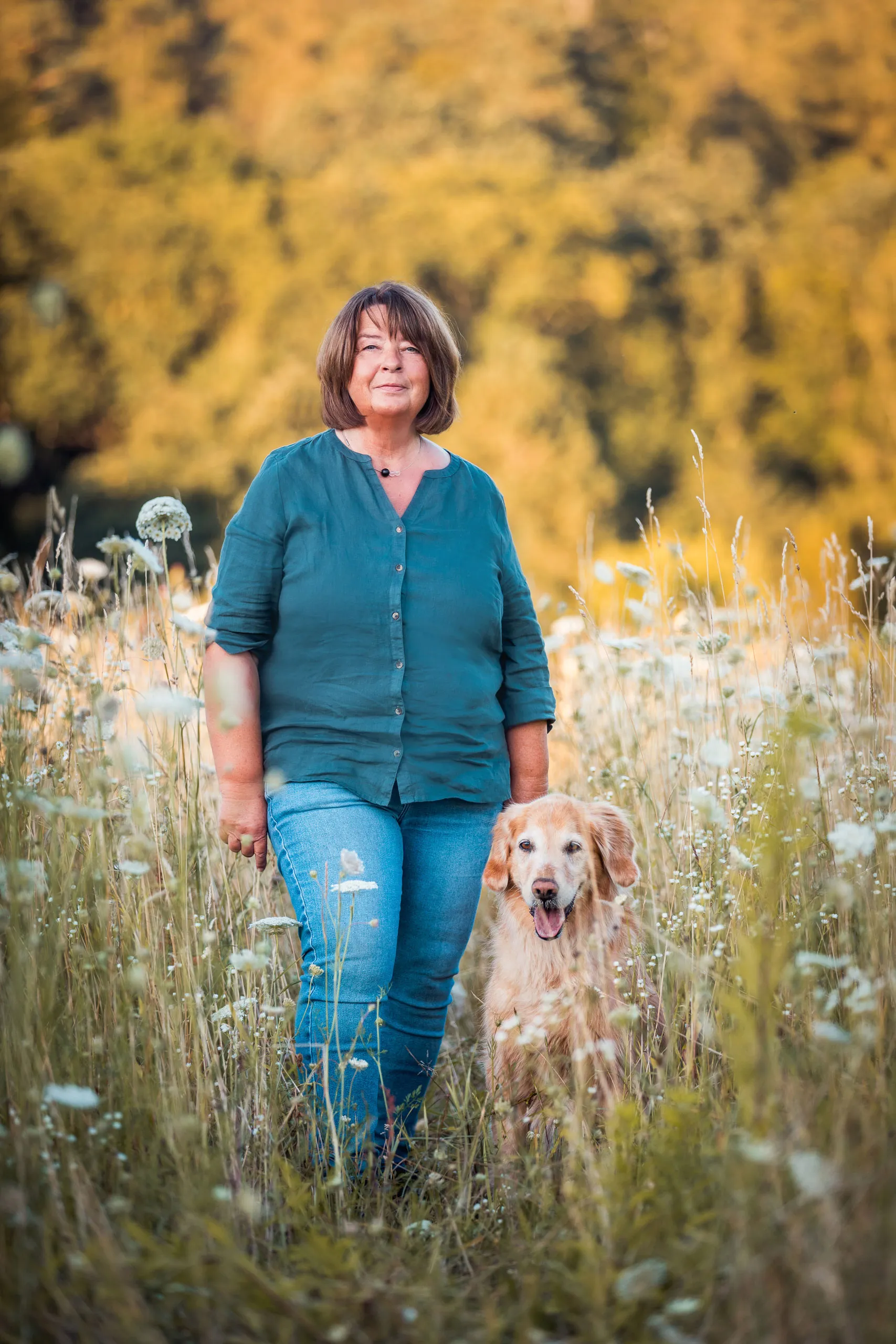 Silvia Hein steht lächelnd auf einer Wiese, ihr Golden Retriever Dori sitzt aufmerksam neben ihr